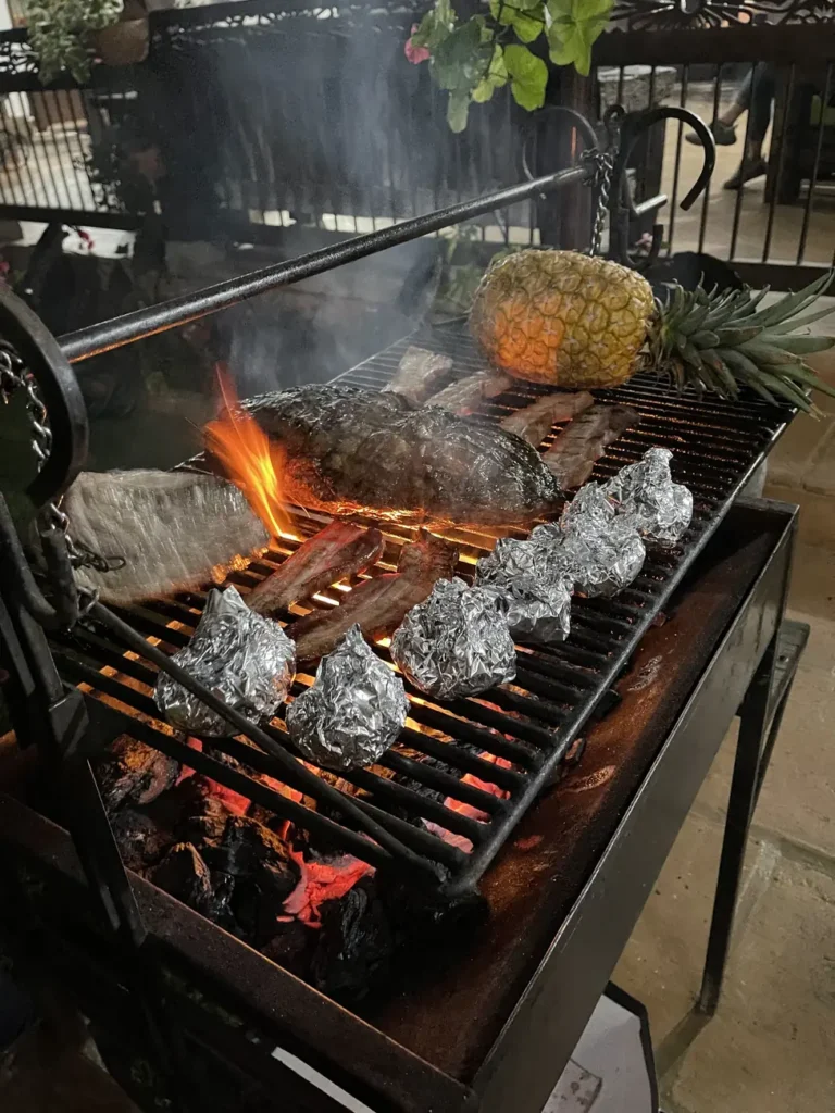 Meat and foil parcels cooking over open coals on an outdoor grill, with a whole pineapple on the rack above