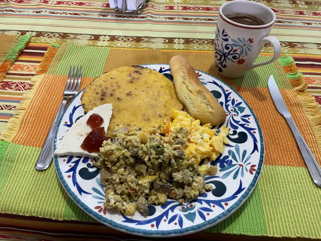 A breakfast plate of eggs, calentado and arepa on a painted ceramic plate beside a cup of coffee