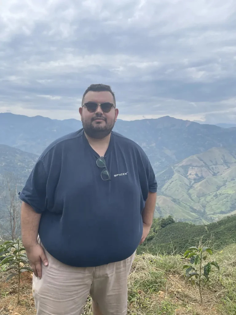 Daniel Molinares standing on a grass ridge with layered coffee covered mountains behind him