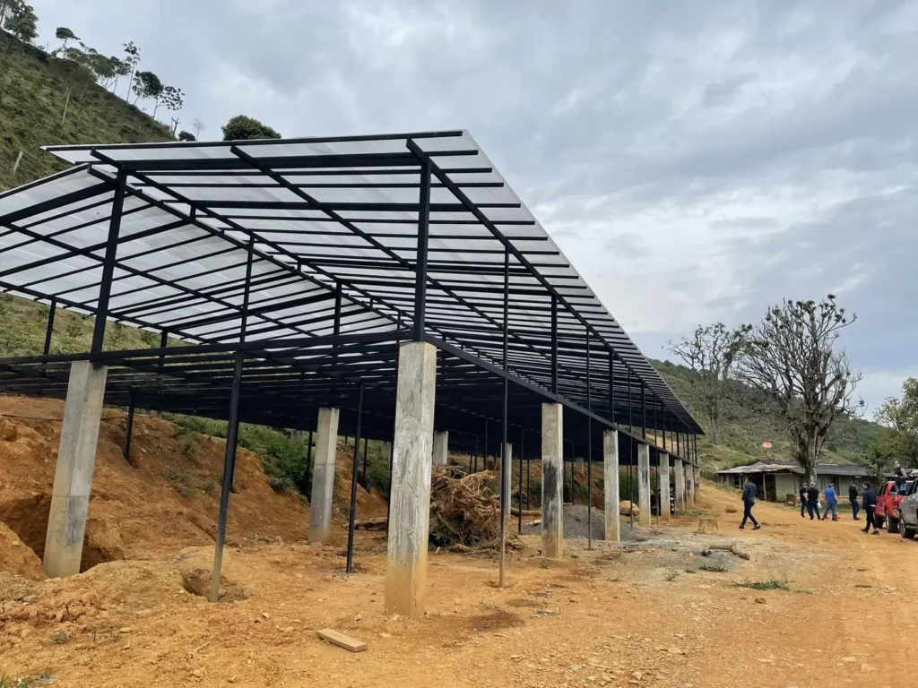 The bare steel frame of a new drying house standing on a red earth cut above the coffee slopes