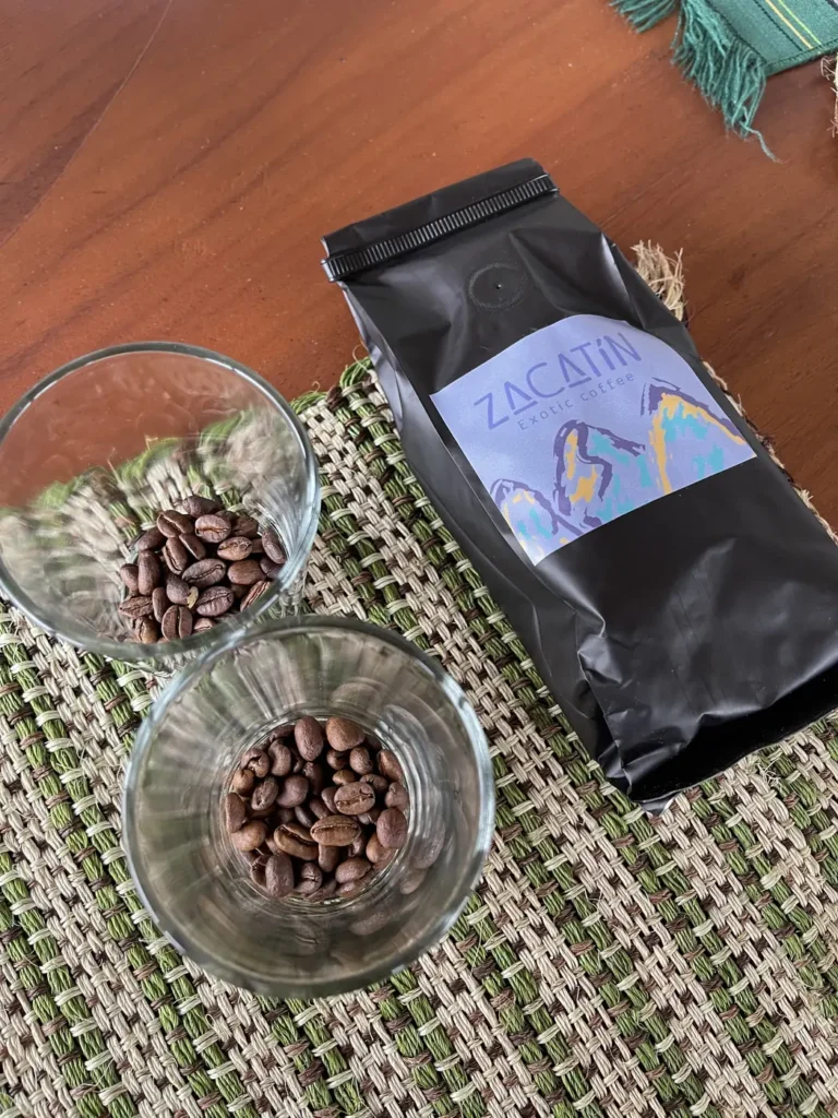 A black El Zacatin coffee bag on a woven mat beside a glass bowl of roasted coffee beans