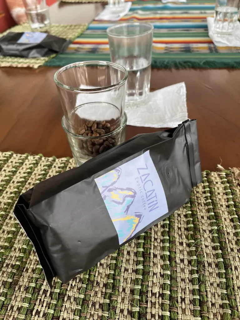 A black El Zacatin coffee bag standing on a woven table mat beside a water glass